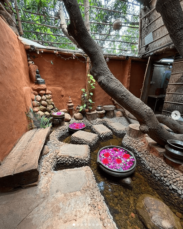 AuraKalari : A serene outdoor setting features a narrow pathway of stepping stones over a shallow pond. Large bowls filled with water and pink flowers are placed on the stones. A tree leans over the courtyard, enhancing the tranquil ambiance, perfect for a nature-inspired vacation. Buddha statues grace the background.