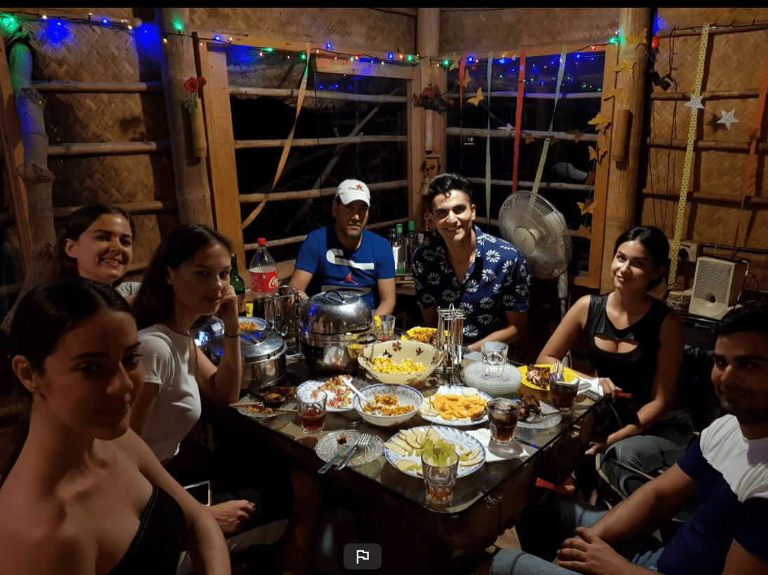 AuraKalari : A group of seven people is seated around a table, adorned with various food items and drinks, in a cozy, wooden setting perfect for aurakalari dining. Festive string lights and hanging decorations create a warm ambiance. Everyone is smiling, clearly enjoying their meal together.