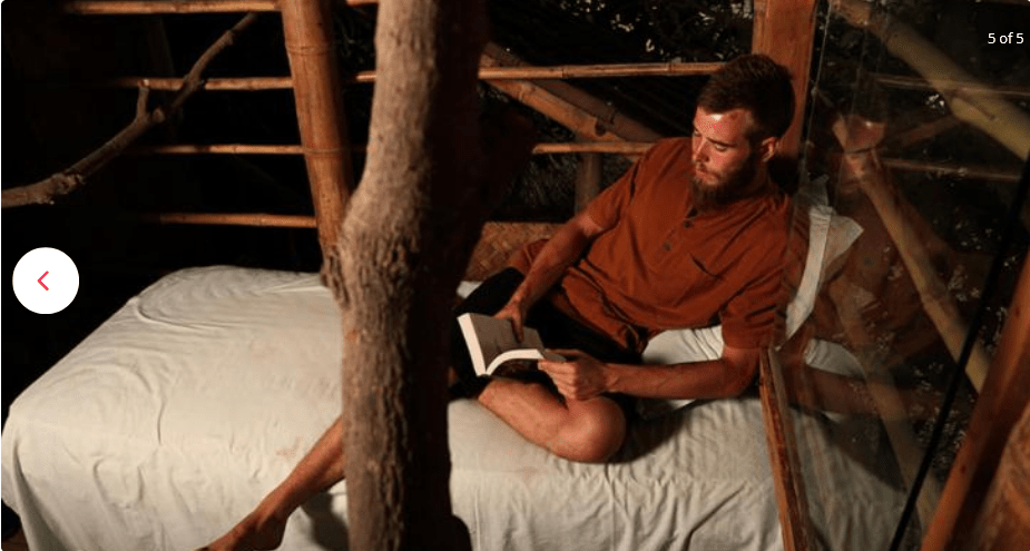 AuraKalari : A man with a beard is lying on a bed made up of white sheets in a rustic mudhouse. He’s reading a book under dim lighting. The bed is situated against a bamboo structure, and there's a large branch in the foreground. Clad in a reddish-brown shirt and dark shorts, he appears immersed in his vacation read.