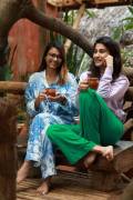 AuraKalari : Two women sit on a wooden bench, both smiling and holding mugs of tea. The woman on the left, an AuraKalari model, is wearing blue and white patterned pajamas and glasses. The woman on the right sports a light purple top with green pants. They are in an outdoor setting with lush plants and wooden structures around them.