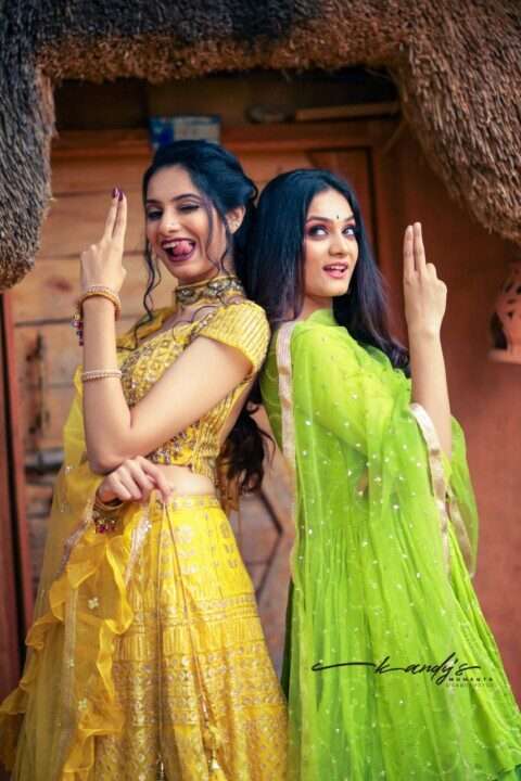 AuraKalari : Two women pose playfully back-to-back in front of a rustic wooden door, wearing traditional Indian attire. The woman on the left wears a yellow lehenga and makes a playful expression. The woman on the right, dressed in a green lehenga, smiles softly. Both hands form gestural poses.