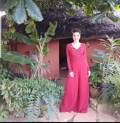 AuraKalari : A woman in a long-sleeved, red dress stands smiling in front of a thatched-roof house surrounded by lush green plants and trees. The house has a rustic feel with a brick exterior and an open doorway. The scene is tranquil, with the woman and natural elements creating a harmonious setting.