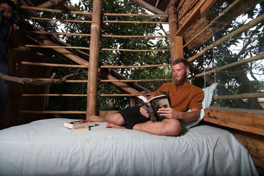 AuraKalari : A man with a beard is sitting cross-legged on a bed inside a rustic, open-walled AuraKalari treehouse, reading a book. He is casually dressed in a brown shirt and black shorts. The treehouse features bamboo poles and large windows offering a view of surrounding trees. Another readbook lies next to him on the bed.