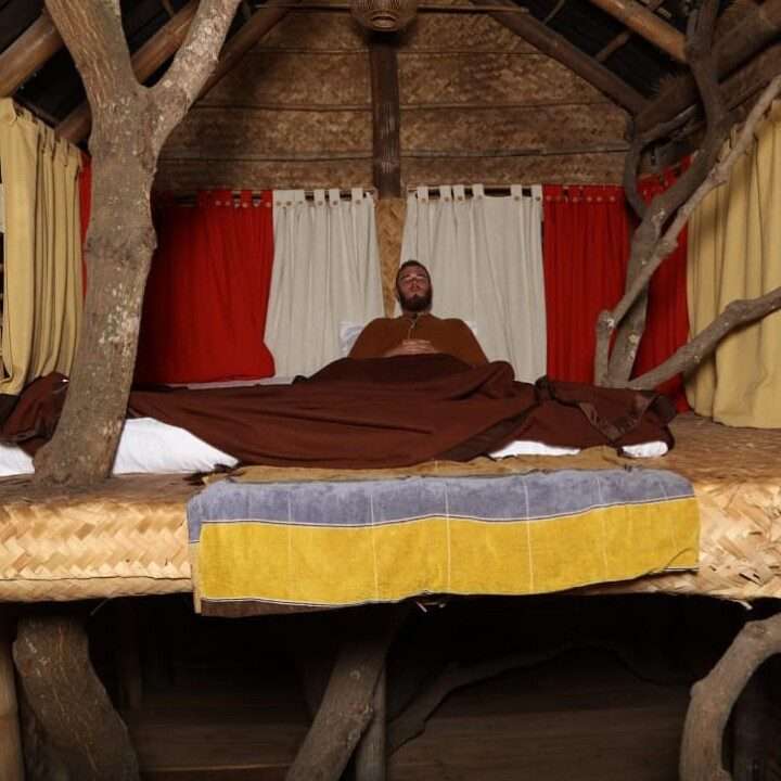AuraKalari : A person covered with a brown blanket lies on a rustic bed inside a treehouse, embracing the ultimate AuraKalari Relax experience. The bed features a woven mat and wooden frame integrated with tree branches. Yellow, gray, and white towels are draped at the foot of the bed as red and yellow curtains create a cozy ambiance.
