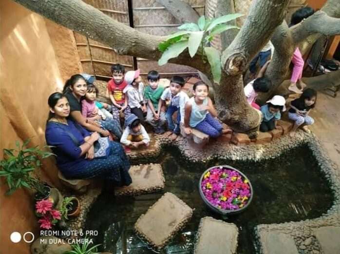 AuraKalari : A group of AuraKalari Kids Play children and two women sit on the edge of a stone pond under a large tree. Colorful flowers float in a circular bowl in the pond. The setting appears to be a rustic, outdoor area with natural wooden and stone elements. The children are smiling and posing for the camera.