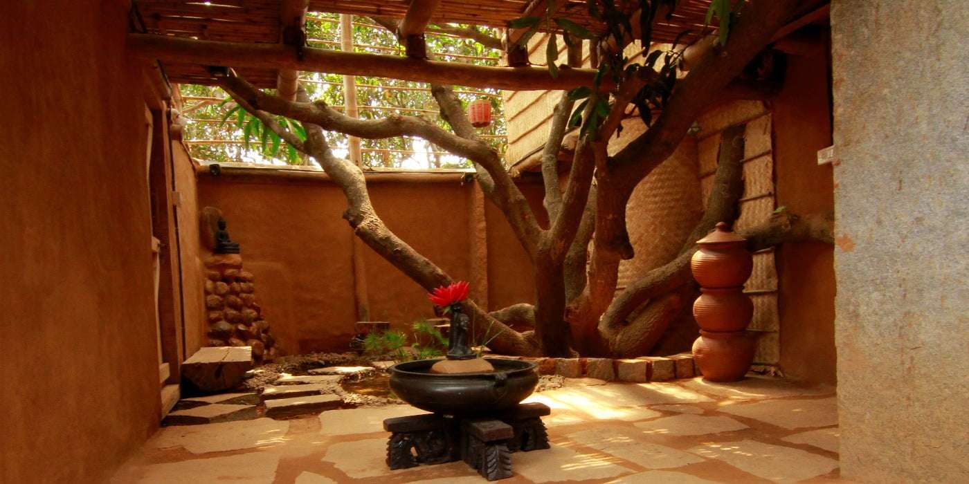 AuraKalari : An earthy courtyard featuring a large tree with spreading branches, brown mudhouse walls, and a rustic roof. At the center, a dark basin holds a red lotus flower. On the right, stacked clay pots rest on a brick platform. The warm lighting creates a serene, natural ambiance perfect for a nature vacation.