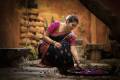 AuraKalari : A woman dressed in a traditional pink and navy blue sari, with her hair tied back and adorned with white flowers, is crouched down, gently placing flowers in a bowl of water. The setting is rustic, with stone walls and a wooden texture, creating a serene and cultural ambiance.